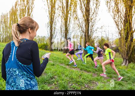 Trainer, der eine Gruppe von Läuferinnen im Freien trainiert, indem er sie bergauf laufen lässt. Stoppuhr in der Hand, Sportbekleidung und professionelle Luft Stockfoto