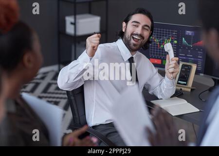 Junger erfolgreicher Makler, der die Aktien telefonisch an den Kunden verkauft, während er mit Kollegen im Büro zusammenarbeitet Stockfoto