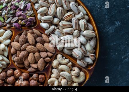 Sortiment von Muttern in Keramikschüssel auf schwarzem Hintergrund, Nahaufnahme, Draufsicht, Kopierbereich. Cashew, Haselnüsse, Pistazien und Mandelnüsse zum Essen. Vegetation Stockfoto