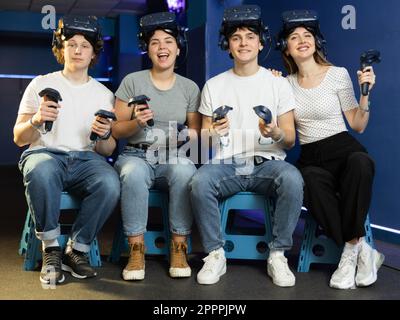 Vier lächelnde Schüler mit Virtual-Reality-Geräten sitzen und posieren im modernen Gaming-Club Stockfoto