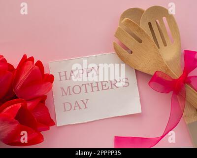 Rote Tulpen blühen mit Küchenutensilien auf rosa Hintergrund mit weißem Papier mit Happy Mütters Day. Grußkarte zum Frauen- und Muttertag. Geburtstag congra Stockfoto