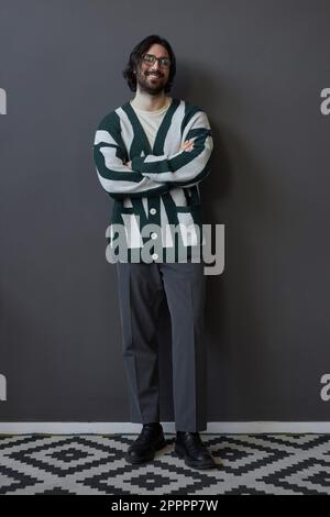 Vertikales Bild eines stilvollen jungen Mannes in Strickjacke, der vor der grauen Wand vor der Kamera lächelt Stockfoto