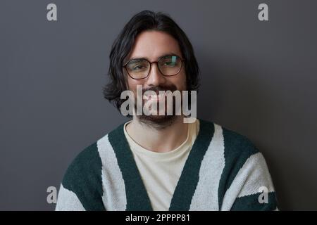 Porträt eines lächelnden gutaussehenden Mannes mit Brille und langen Haaren, die vor grauem Hintergrund vor der Kamera lächeln Stockfoto