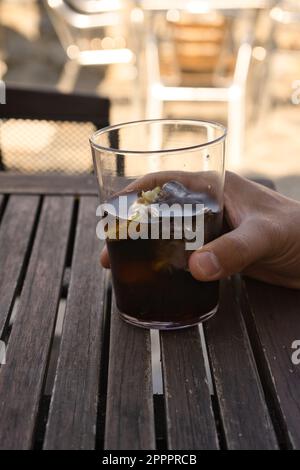 Männliche Hand hält ein Cola-Getränk in einem Kristallglas auf einem Holztisch. Im Freien. Stockfoto