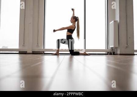 Starke weiße Frau, die mit einem Knie auf Yogamatte steht und Kurzhanteln für Armübungen benutzt. Gut ausgebildete Dame in schwarzer Aktivkleidung, die in der Nähe von Panoramafenstern mit Blick auf die Stadt trainiert. Stockfoto