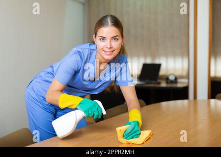 Junge weibliche Hausmeisterin Staub Möbel im Büro mit Reinigungsgeräten Stockfoto