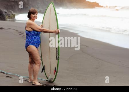 Eine freudige Erwachsene Frau mittleren Alters im Badeanzug hält ein Surfbrett zum Surfen, genießt den Sommerurlaub am Strand am Meer. Aktiver Lebensstil, extrime Stockfoto