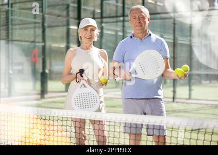 Alter Mann und Frau posieren mit Padel Schläger in den Händen neben dem Netz auf dem Tennisplatz Stockfoto