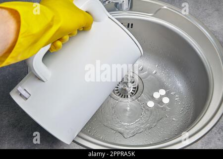 Eine Person in gelben Gummihandschuhen gießt heißes Wasser mit Dampf aus einem elektrischen Wasserkocher in ein Metallwaschbecken Stockfoto