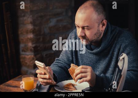 Ein Mann isst ein Sandwich und sieht zu Hause auf das Telefon. Ein gewöhnlicher Mann mittleren Alters. Essen mit einem Telefon in der Hand. Stockfoto