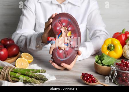 Frau, die ein Nierenmodell in der Nähe verschiedener Produkte hält, auf einem weißen Holztisch, Nahaufnahme Stockfoto