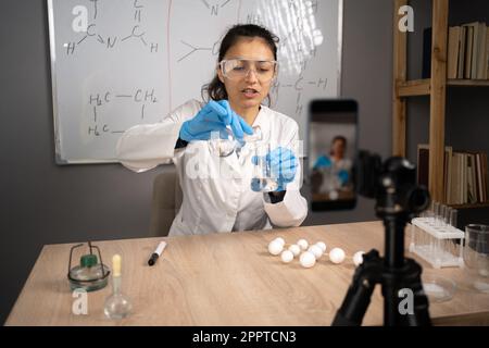 Online-Lehrer, Mentor. Klassenzimmer für Schulwissenschaften. Der Lehrer erklärt Chemie. Junge Frau unterrichtet und nimmt Videokurs auf, Smartphone auf einem Stativ Stockfoto