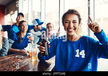 Wir haben den ersten Platz nach Hause genommen. Porträt einer Frau, die einen Finger hochhält, während sie sich mit Freunden in einer Bar ein Sportspiel ansieht. Stockfoto