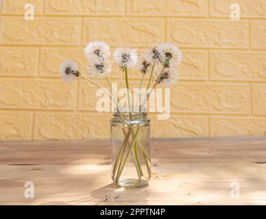 Ein Blumenstrauß Löwenzahn in einem transparenten Glasgefäß auf einem Holztisch mit Ziegelhintergrund Stockfoto