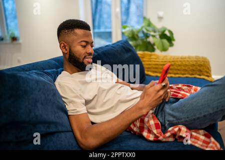 Ein entspannter lächelnder afroamerikanischer Mann sitzt auf der Couch und sieht zu Hause auf ein Smartphone. Stockfoto