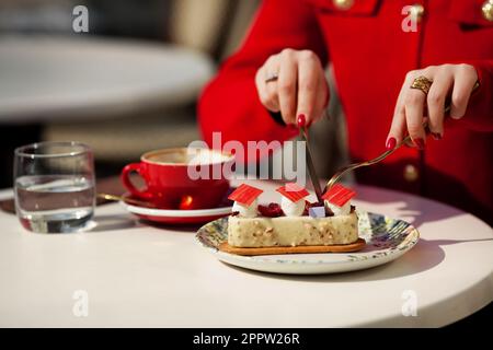 Eine Frau in roten Kleidern isst Kuchen und trinkt Kaffee im Café. Nahaufnahme weiblicher Hände mit Messer und Gabel schneidendem Dessert Stockfoto