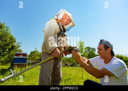 Ein Techniker mit verbandelten Handverletzungen repariert den Trimmer, stellt die Kopfspule des Trimmers ein und hilft seinem Freund im Schutzanzug beim Rasenmähen Stockfoto