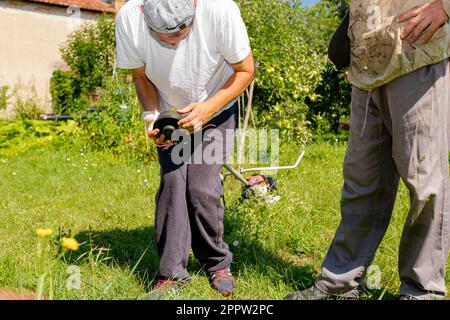 Ein Techniker mit verbandelten Handverletzungen repariert den Trimmer, stellt die Kopfspule des Trimmers ein und hilft seinem Freund im Schutzanzug beim Rasenmähen Stockfoto