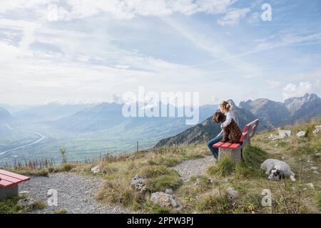 Ein Mädchen mit Hunden sitzt auf einer Bank und genießt die malerische, majestätische Aussicht auf die Bergkette in der Schweiz Stockfoto