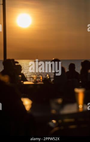 Abgeschirmte Leute, die Bier auf der Terrasse am Wasser trinken, mit Blick auf das Meer bei Sonnenuntergang Stockfoto