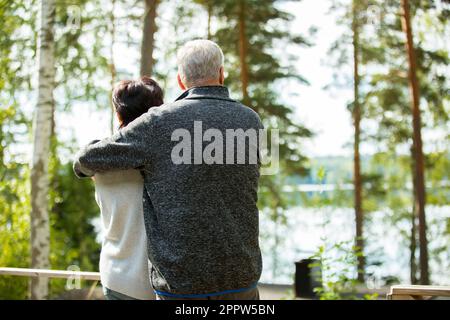 Reifes Paar, das auf der Veranda steht und sich umarmt. Sie lächeln und schauen auf die wunderschöne Aussicht - Wald und See. Glückliches Seniorenpaar umarmt sich Stockfoto