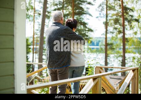 Reifes Paar, das auf der Veranda steht und sich umarmt. Sie lächeln und schauen auf die wunderschöne Aussicht - Wald und See. Glückliches Seniorenpaar umarmt sich Stockfoto