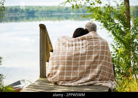 Mature couple sitting on wooden steps and hugging. They are wrapped in blanket, smiling and looking at beautiful view - forest and lake. Happy senior Stockfoto