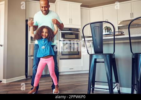 Zuhause ist der Ort, an dem der Spaß stattfindet. Ein glückliches kleines Mädchen, das auf den Füßen ihres Vaters balanciert, während sie zusammen in der Küche herumlaufen. Stockfoto