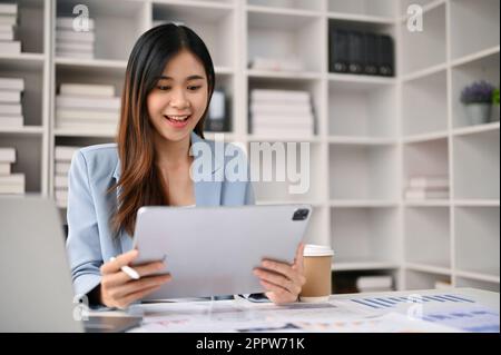 Charmante und lächelnde asiatische Millennials-Geschäftsfrau oder Büroangestellte, die ihr digitales Tablet an ihrem Schreibtisch im Büro verwendet. Stockfoto