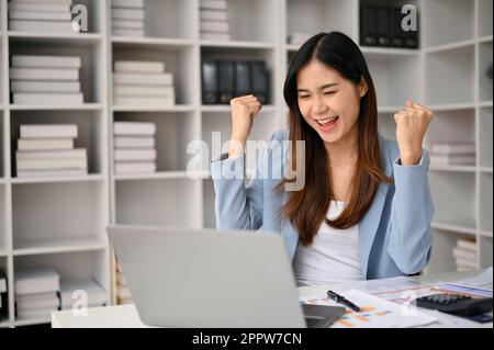 Aufgeregte und fröhliche asiatische Geschäftsfrau, die die Hände hebt, geballte Fäuste zeigt, ihren beruflichen Erfolg feiert und freut, empfängt Stockfoto