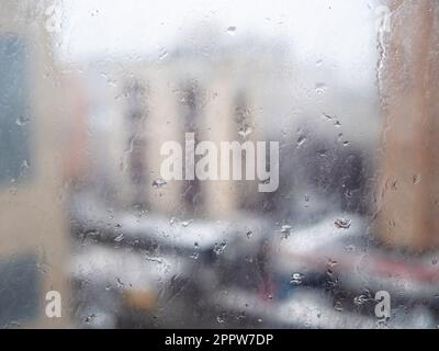 Kalte Regentropfen auf Fensterglas in der Wohnung der Stadt und Blick auf Stadthäuser im Hintergrund Stockfoto