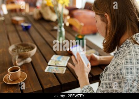 Eine schöne junge Frau rät auf Karten mit Tarot, Runen auf einem Holztisch und benutzt eine Online-App im Telefon, um die Verwirrung zu interpretieren, die Inneneinrichtung, Ca Stockfoto