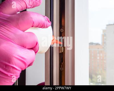 Hand in rosafarbenem Handschuh schmiert die Schließung des Fensterrahmens in der Wohnung eines mehrstöckigen Stadthauses aus der Ölkanne Stockfoto