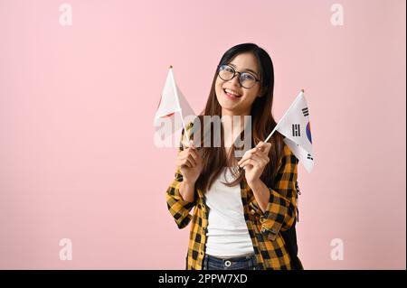 Lächelnde und positive junge asiatische College-Studentin mit der Flagge von Korea und Japan vor pinkfarbenem isoliertem Hintergrund. Im Ausland studieren und tauschen Stockfoto