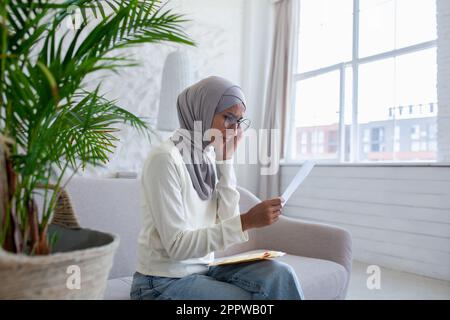 Muslimische Frau zu Hause in Hidschab-Aufregung, arabische Frau erhielt E-Mail-Benachrichtigung mit schlechten Nachrichten, Frau liest nachdenklich und besorgt auf dem Sofa im Wohnzimmer. Stockfoto