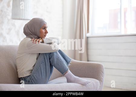 Junge Frau, die sich in Depressionen aufregte, sitzt zu Hause auf dem Sofa, muslimische Frau in Hijab, dicht im Wohnzimmer. Stockfoto