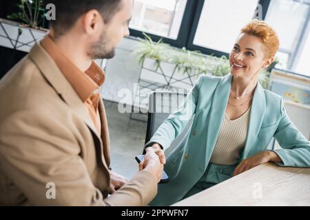 Freudige rothaarige Geschäftsfrau, die sich die Hand schüttelt mit einem verschwommenen Partner im Büro, Börsenbild Stockfoto