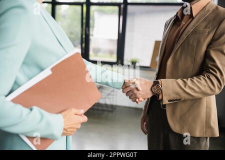 Zugeschnittene Ansicht einer Geschäftsfrau mit Ordner, der einem Partner im Büro die Hand schüttelt, Aktienbild Stockfoto
