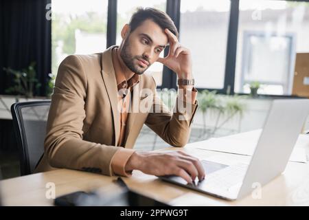 Gelangweilter Geschäftsmann in beigefarbenem Blazer, der auf ein Notebook schaut, während er im Büro am Arbeitsplatz sitzt, Bild von Stock Stockfoto