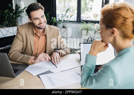 Lächelnder, bärtiger Manager, der auf Karten in der Nähe einer Geschäftsfrau zeigt, auf verschwommenen Vordergrund im Büro, Aktienbild Stockfoto