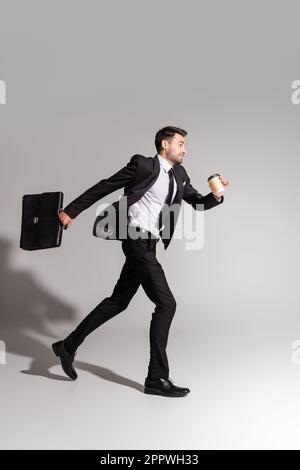 Seitenansicht eines positiven Geschäftsmanns im schwarzen Anzug mit Aktentasche und Pappbecher auf grauem Hintergrund, Aktienbild Stockfoto