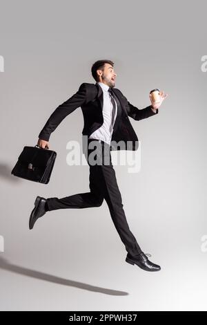 Seitenansicht eines begeisterten Geschäftsmanns in schwarzem Anzug mit Lederaktentasche und Pappbecher auf grauem Hintergrund, Stockbild Stockfoto