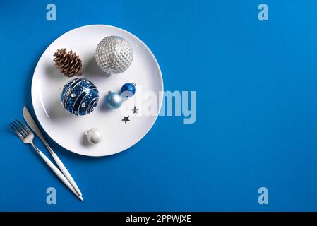 Kreative Tischgestaltung Zu Weihnachten. Festliche Ornamente und Einrichtung auf weißem Teller auf blauem Hintergrund, Blick von oben, Kopierbereich. Weihnachtsessen, Partydesign Stockfoto