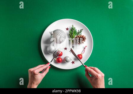 Kreative Tischeinstellung und Frauenhände. Festliche silberne Kugeln und Kiefernzapfen auf weißem Schild auf grünem Hintergrund, flach liegend, Kopierbereich. Essen Sie Stockfoto