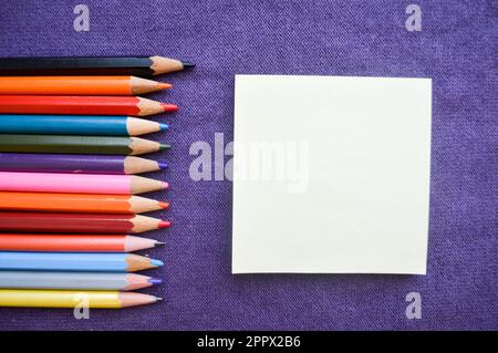 Mehrfarbige, leuchtende, farbenfrohe Bleistifte zum Zeichnen auf der linken Seite und ein Notizblock auf der rechten Seite für Ihren Text auf einem lilafarbenen Texthintergrund. Stockfoto