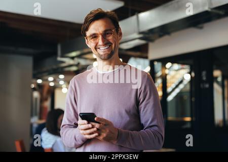 Porträt eines glücklichen Geschäftsmanns, der ein Handy in einem Büro hält. Professioneller Geschäftsmann, der auf die Kamera schaut, während er am Arbeitsplatz steht. Stockfoto