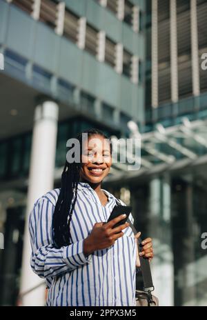 Glückliche schwarze Geschäftsfrau, die auf dem Weg zur Arbeit in der Stadt vor der Kamera lächelt. Porträt einer fröhlichen jungen Geschäftsfrau, die ein Smartphone benutzt, während sie Stockfoto