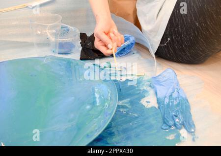 Der Prozess, ein hausgemachtes, modernes, abstraktes Muster zu kreieren, das mit einem Pinsel aus mehrfarbigem Acrylharz auf einem runden Holzbrett bemalt wurde. Stockfoto