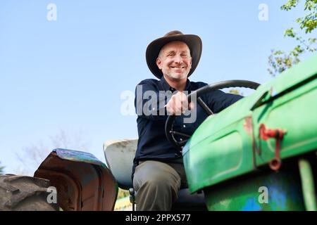 Porträt eines reifen männlichen Landwirts, der am sonnigen Tag einen Traktor mit klarem Himmel im Hintergrund fährt Stockfoto