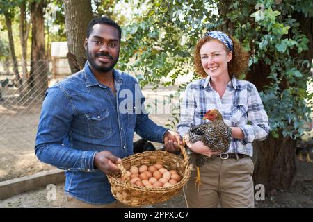 Porträt von lächelnden multikulturellen männlichen und weiblichen Landwirten mit Hühnern und Eiern im Geflügelzuchtbetrieb Stockfoto